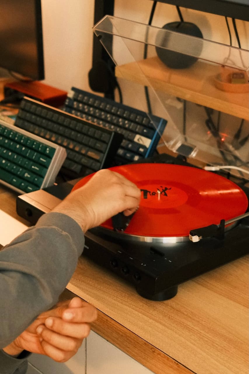 Hand placing red vinyl record on turntable with mechanical keyboards and headphones on wooden desk
